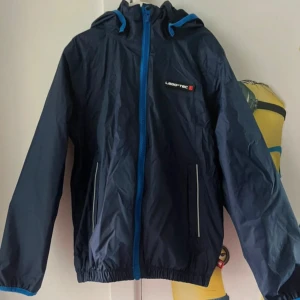 Mörkblå vindjacka från LEGO Wear - Snygg mörkblå vindjacka från LEGO Wear med huva och blå detaljer vid dragkedja och ärmslut. Jackan är vattentät, har tejpade sömmar och två fickor med reflexdetaljer. Perfekt för blåsiga och regniga dagar, andas bra och har elastisk nederkant.