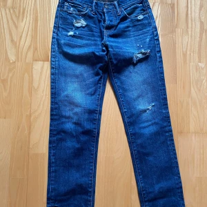 Blå jeans Abercrombie & Fitch W28 L30 - Snygga blå jeans från Abercrombie & Fitch med slitna detaljer och hål på både fram- och baksida. Klassisk femficksmodell med raka ben och normal passform. Jeansen har orangea sömmar och är tillverkade i jeansmaterial. Perfekt för dig som gillar en avslappnad och trendig look.