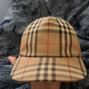 Burberry rutig keps beige M - Snygg Burberry keps i klassiskt beige rutigt mönster med svarta, röda och bruna linjer. Märke med 'BURBERRY LONDON ENGLAND' baktill. Justerbar snapback och tillverkad i bomull och polyester. Perfekt för dig som vill ha en ikonisk streetstil. För bara 1700kr MALMÖ!!!!! ÄKTA!!!!
