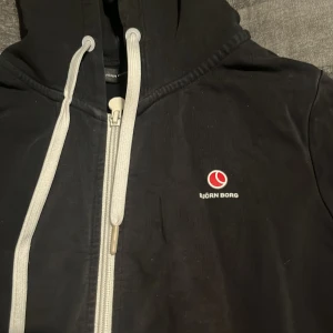 Svart hoodie från Björn Borg med dragkedja - Svart hoodie från Björn Borg med vit dragkedja och vita snören i huvan. Liten röd och vit logga på bröstet. Tröjan har långa ärmar, ribbade muddar och två fickor framtill. Perfekt för chill eller träning. 900 kr ny. 
