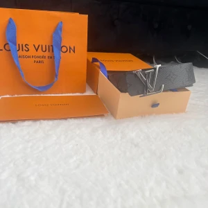 Svart Louis Vuitton skärp med LV-spänne - Säljer ett stilrent svart Louis Vuitton bälte med ikoniskt LV-spänne i silver. Bältet är tillverkat i präglat skinn med diskret monogrammönster. Perfekt accessoar för att lyfta din outfit med lyxig känsla och snygg finish. Helt ny oanvänt 