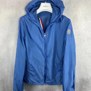 Blå vindjacka från Moncler - Snygg blå vindjacka från Moncler med huva och dragkedja framtill. Jackan har Moncler-logga på ärmen och coola detaljer med röd och vit rand på insidan av kragen. Perfekt för dig som gillar sportig och stilren look.