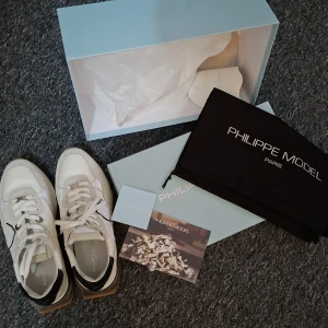 Philippe Model vita sneakers strl 40 - Snygga vita sneakers från Philippe Model med beige mockadetaljer, svart häl och gummisula med transparenta inslag. Skorna har snörning, rund tå och coolt sköldmärke på sidan. Perfekt för dig som gillar stilrena och trendiga sneakers med lyxig känsla. Bara använda 1 gång. Säljer dom då de är för små för mig. Priset går att diskutera vid snabb affär! Känn dig fri att höra av dig vid minsta fundering! 😁 kartong och allt medföljer! 