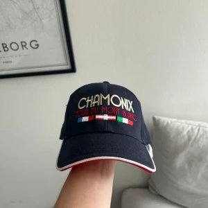 Mörkblå Chamonix keps med brodyr - Snygg mörkblå keps med broderad text 'CHAMONIX TOUR DU MONT BLANC' och flaggor i vitt, rött, blått och grönt framtill. Kepsen har böjd skärm med vit kant och röd undersida samt justerbar rem bak. Perfekt för dig som gillar friluftsliv och alp-vibes.
