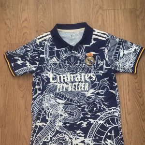Real Madrid drakmönstrad fotbollströja - Säljer en Real Madrid fotbollströja från Adidas i marinblått med vit drakmönster över hela tröjan. Tröjan har korta ärmar, krage och gula detaljer vid ärmslut. Materialet är lätt och andas, perfekt för träning eller match. Broderat klubbmärke på bröstet.