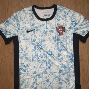 Portugal Nike kortärmad fotbollströja - Snygg Portugal fotbollströja från Nike med unikt blått mönster på vit bakgrund och svarta detaljer vid krage och sidor. Tröjan har broderat Portugal-emblem på bröstet och är tillverkad i lätt, ventilerande material. Perfekt för dig som vill sticka ut på planen eller läktaren.