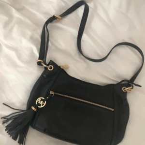 Svart axelväska Michael Kors - Snygg svart axelväska från Michael Kors i skinn med guldfärgade detaljer och en dekorativ tofs med MK-logga. Väskan har justerbar rem, dragkedja framtill