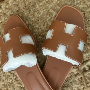 Bruna sandaler med H-design - Snygga bruna sandaler med öppen tå och platt sula. De har en stilren H-formad rem i skinn med vita sömmar som ger en exklusiv vibe. Perfekta för varma dagar och passar till både jeans och klänning.(passar fötter som är 25 cm och det motsvarar storlek 39)