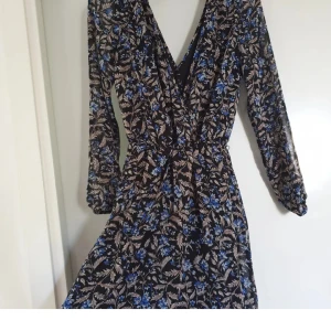 Mörkblå blommig kortklänning med v-ringning - Mörkblå kortklänning med v-ringad omlottfront och långa ärmar. Klänningen har ett blommigt mönster i blått och beige, med lätt transparent material och resår vid ärmsluten. Använd 2 ggr.