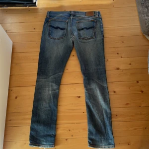 Blå Nudie Jeans straight fit - Säljer ett par klassiska blå jeans från Nudie Jeans med snyggt slitna detaljer och orange sömmar. Modellen har raka ben och fem fickor, med Nudie-logga på bakfickan och läderpatch i midjan. Jeansen är tillverkade i bomull och har en cool faded look.