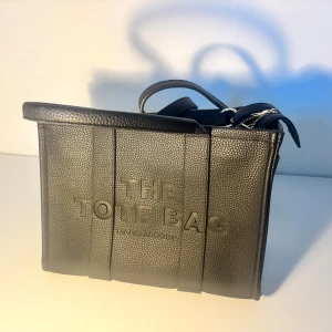 Svart Tote Bag från Marc Jacobs - Snygg svart Tote Bag från Marc Jacobs i präglat skinn med tydlig logga framtill. Väskan har rejäla handtag, guldfärgade detaljer och dragkedja upptill. Perfekt storlek för att få plats med allt du behöver och stilren design som sticker ut. I don’t know if it’s real 