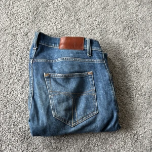 Blå jeans Pistolero 33/34 - Säljer ett par klassiska blå jeans från Pistolero i storlek 33/34. Jeansen har en rak passform med snygga slitningar och kontrastsömmar. De är tillverkade i slitstarkt denim och har en läderpatch bak i midjan. Perfekt för dig som gillar en tidlös stil.