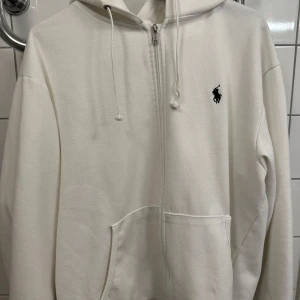 Vit hoodie från Polo Ralph Lauren - Vit hoodie från Polo Ralph Lauren med dragkedja framtill och klassisk svart logobrodyr på bröstet. Tröjan har huva med snörning, känguruficka och långa ärmar. Mjuk bomullskvalitet som är skön att ha på sig. Perfekt för chill dagar och streetwear.