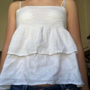 Vit somrig volangtopp med broderade blommor – stl 170 - Söt vit topp med tunna axelband, volang och broderade blommor. Luftigt och lätt material – perfekt för sommaren! Passar både till vardags och festligare tillfällen. Storlek 170 och i mycket fint skick, knappt använd.