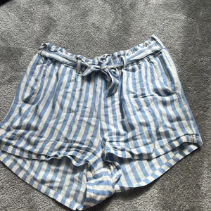 Randiga shorts med knyt från KappAhl - Snygga shorts från KappAhl i ljusblått och vitt med vertikala ränder. Modellen har hög midja, praktiska fickor och ett matchande knytband i midjan. Materialet är mjukt och känns som bomull, perfekt för varma dagar. Passformen är loose och avslappnad.