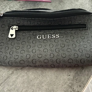 Svart magväska från Guess - Snygg svart magväska från Guess med präglat mönster av små G:n över hela väskan. Väskan har en dragkedja framtill med metall-detalj och logotypen GUESS i silver på framsidan. Perfekt för att bära det viktigaste och ha händerna fria. Köparen betalar frakt. 