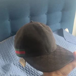 Svart Gucci keps med GG-mönster - Snygg svart keps från Gucci med klassiskt GG-monogram över hela kepsen. På sidorna finns de ikoniska gröna och röda ränderna. Justerbar rem baktill och böjd skärm. Tillverkad i bomullsmix med exklusiv känsla – perfekt för dig som vill sticka ut.