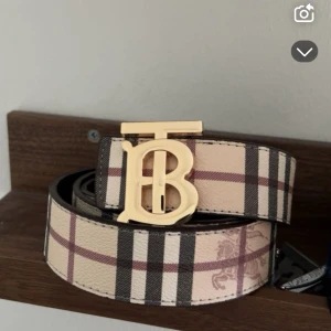 Burberry bälte - Snyggt Burberry bälte med klassiskt rutigt mönster i beige, brun och svart. Bältet har en stor guldfärgad spänne med TB-logga framtill. Tillverkat i skinn med exklusiv känsla och ikonisk design som sticker ut.