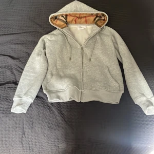 Grå hoodie från Burberry med dragkedja - Säljer en grå hoodie från Burberry med klassiskt rutigt foder i huvan och broderad logga på bröstet. Tröjan har snörning i huvan, ribbade muddar och dragkedja framtill. Perfekt för dig som vill ha en stilren och lyxig look.