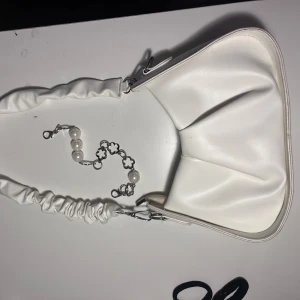 Vit axelväska med extra band - Snygg vit axelväska med ett axelband med mjuk, rynkad design. Väskan har en dragkedja upptill och silverfärgade metalldetaljer. Perfekt storlek för mobil, plånbok och småsaker. Stilren look som lyfter din outfit. Det kommer med justerbart band som kan göra det längre! I mycket bra skick!