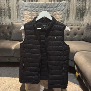 Svart quiltad dunväst Lyle & Scott - Svart dunväst från Lyle & Scott Junior med gul logotyp på bröstet. Västen har quiltad design, två sidofickor med dragkedja och hög krage. Perfekt för lager-på-lager och snygg till hoodie eller t-shirt.