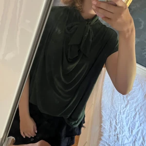 Grön blus med knyt från By Malene Birger - Snygg mörkgrön blus från By Malene Birger med korta ärmar och ett elegant knyt i halsen. Blusen har ett mjukt, draperat fall och är tillverkad i ett tunt, skönt material som känns lyxigt mot huden. Perfekt för dig som gillar stilrena detaljer.