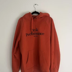 Orange hoodie från Peak Performance - Säljer en orange hoodie från Peak Performance med svart tryck på bröstet. Tröjan har huva med snörning, stor magficka och ribbade muddar. Perfekt för chill dagar och har en loose passform. Materialet är mjuk bomull som känns skönt mot huden.