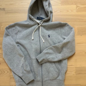 Grå hoodie från Polo Ralph Lauren - Snygg grå hoodie från Polo Ralph Lauren med dragkedja och vit snörning i huvan. Klassisk logga broderad på bröstet i mörkblått. Mjuk bomullskvalitet, känguruficka framtill och ribbade muddar vid ärmar och nederkant. Perfekt för chill dagar.
