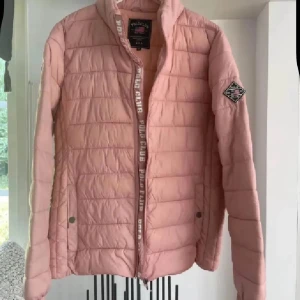 Rosa jacka  från Polo Club - Snygg ljusrosa pufferjacka från Polo Club med quiltad design och hög krage. Jackan har dragkedja framtill, två fickor med tryckknappar och en cool patch på ärmen. Perfekt för dig som vill ha en stilren och bekväm jacka med sportig vibe.