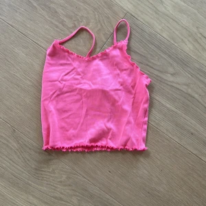 Rosa croppad linnetopp Gina Tricot - Säljer en ljusrosa croppad linnetopp från Gina Tricot med smala axelband och vågig kant nertill. Toppen är ärmlös och gjord i mjuk bomull, perfekt för varma dagar. Enkel och snygg design som ger en chill vibe.