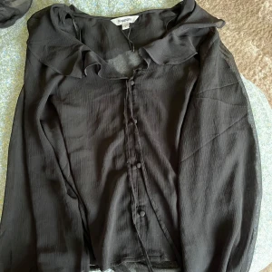 Svart blus med volang från Kappahl - Snygg svart blus från Kappahl med volang vid kragen och knappar framtill. Lätt transparent material och långärmad modell som ger en cool och elegant vibe. Hör av dig vid frågor!💕