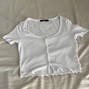 Vit ribbad croppad topp från Bik Bok - Säljer en vit croppad topp från Bik Bok med ribbad struktur och korta ärmar. Toppen har rundad hals, knappar framtill och vågiga kanter vid ärmslut och nederkant. Perfekt för dig som gillar en enkel men söt stil.