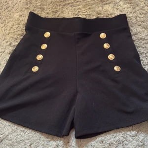 Svarta högmidjade shorts med guldknappar H&M - Snygga svarta shorts från H&M med hög midja och sex dekorativa guldknappar framtill. Klassisk rak passform och mjukt, lite stretchigt material som sitter bekvämt. Perfekt för dig som gillar en stilren look med coola detaljer.