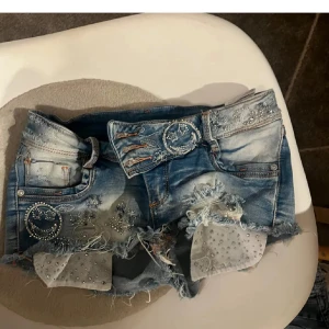 Blå jeansshorts med pärlor och slits - Snygga blå jeansshorts från Angels i storlek S med slitna detaljer, fransig kant och coola pärlbroderier framtill. Shortsens midja har dubbla knappar och dekorativa sömmar. Insidan av benen har vita tygpaneler med pärlor för extra bling. Storlek xs, köp ej direkt!❤️