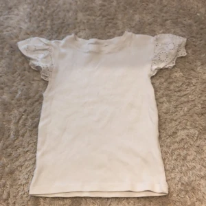 Vit topp med spetsärmar Gina Tricot - Supersöt vit topp från Gina Tricot i storlek 134/140. Toppen har korta ärmar med volang och spetsdetaljer som ger en extra touch. Materialet är mjukt och bekvämt, perfekt för dig som gillar en clean och fräsch look med lite extra detaljer på ärmarna.