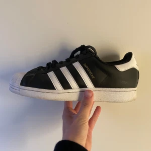 Adidas Superstars - Säljer dessa Adidas Superstars sneakers i svart och vitt med de ikoniska tre ränderna på sidorna och guldiga detaljer på plösen. Skorna har snörning, rund tå och platt sula. Har vissa skavanker & snörena är dammiga av att ha suttit i källaren ett bra tag, men både av det försvinner i en tvätt eller två!