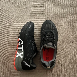 Adidas Terrex GTX svarta sneakers - Adidas Terrex GTX sneakers i svart med grå och orange detaljer. Skorna har grov Continental-sula för extra grepp, snörning och GTX-membran som håller fötterna torra. Perfekta för äventyr utomhus och träning. Snygg design med vita logotyper på sidan.