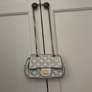 Quiltad silver axelväska Milk x Soda - Snygg quiltad axelväska i metallic silver från Milk x Soda. Väskan har en kedja i guld, vridlås framtill och ett klassiskt rutmönster. Perfekt storlek för det viktigaste och ger en cool touch till din outfit.