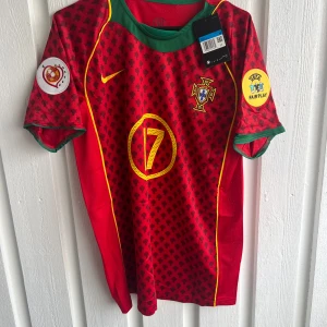 Portugal C. Ronaldo #17 fotbollströja Nike - Snygg Portugal fotbollströja från Nike med C. Ronaldo och nummer 17 i gult på ryggen. Röd bas med svarta mönster, gula detaljer och gröna muddar. Kortärmad modell med officiella UEFA Fair Play och Portugal-emblem. Perfekt för dig som älskar fotboll och Ronaldo.