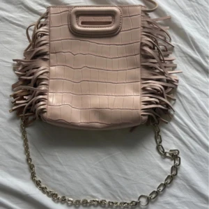 Beige/ljusrosa axelväska med fransar från Maje - Säljer en beige axelväska från Maje med croco-mönstrad yta och snygga fransar på sidorna. Väskan har både axelrem och en cool kedja, samt ett praktiskt handtag upptill. Perfekt för dig som gillar detaljer och stilren design.