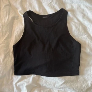 Svart ribbad croppad linnetopp - Svart croppad linnetopp från Shein med ribbad struktur och hög hals. Toppen är ärmlös och har en sportig, clean look som sitter tight mot kroppen. Perfekt att styla med jeans eller shorts för en chill vibe.