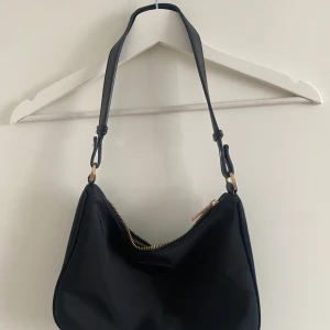 Svart axelväska med dragkedja - Snygg svart axelväska med justerbar rem och guldfärgad dragkedja. Väskan har en mjuk, rundad form och är tillverkad i ett slätt syntetmaterial. Perfekt storlek för att få plats med det viktigaste och passar till många olika stilar.