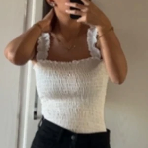 Vit smockad linnetopp med volang - Supersöt vit linnetopp med smockad stretch och volangdetaljer på axelbanden. Toppen är ärmlös och har en croppad passform som passar perfekt till höga jeans. Materialet känns lätt och luftigt, perfekt för varma dagar.