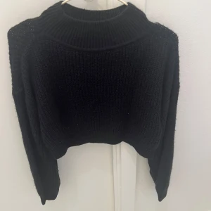 Svart croppad stickad tröja - Säljer en svart croppad stickad tröja med bred ribbad krage och långa ärmar. Perfekt för dig som gillar en chill och trendig look. Tröjan har en loose passform och passar bra till både jeans och kjol. Enkel att styla och supermysig under kyliga dagar.