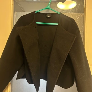 Svart kappa från Gina Tricot - Svart kappa från Gina Tricot i storlek L. Jackan har en stilren och rak design med öppen front och breda slag. Materialet känns mjukt och har en ullig look som passar perfekt för kyliga dagar. Enkel och snygg att matcha med olika outfits.
