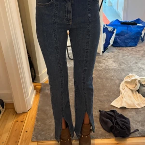 Blå jeans med slits och hög midja - Säljer ett par blå jeans med coola slitsar framtill vid bensluten. De har hög midja och raka ben med markerade sömmar längs framsidan. Jeansen är i klassisk denim och har en snygg, modern look som passar till boots eller sneakers.