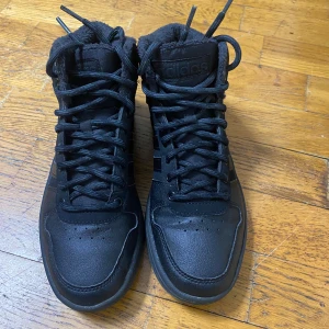 Svarta höga sneakers från Adidas - Det är ett par svarta Adidas högskor med snörning och vad som verkar vara ett varmt foder, perfekt för kallare väder.