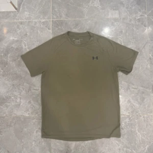Olivgrön t-shirt från Under Armour - Snygg olivgrön t-shirt från Under Armour med diskret logga på bröstet. Modellen är klassisk med rund hals och korta ärmar, perfekt för träning eller chill. Materialet är lätt och andas, vilket gör den skön att bära även när du svettas.