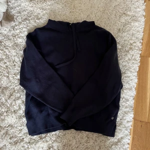 Mörkblå stickad hoodie - Mysig mörkblå hoodie med huva och snörning framtill. Tröjan är stickad och har långa ärmar, perfekt för kyliga dagar. Stilren design utan mönster och med en liten metallknapp vid nederkanten. Passar dig som gillar enkel och clean look.