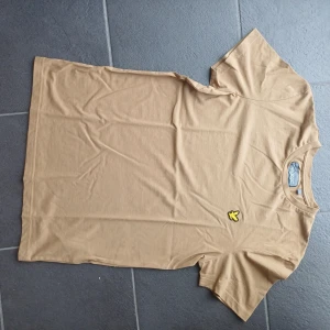 Beige t-shirt från Lyle & Scott - Snygg beige t-shirt från Lyle & Scott i mjuk bomull. Klassisk passform med korta ärmar och rund hals. På bröstet sitter den ikoniska gula fågel-loggan som ger en clean och stilren vibe. Perfekt för dig som gillar enkel och tidlös stil.
